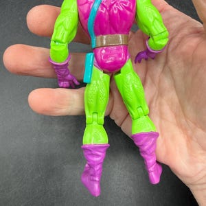 May include: A Green Goblin action figure with a purple head, gloves, and boots. The figure has a pink torso with a brown belt and a blue strap. The figure is held in a hand.