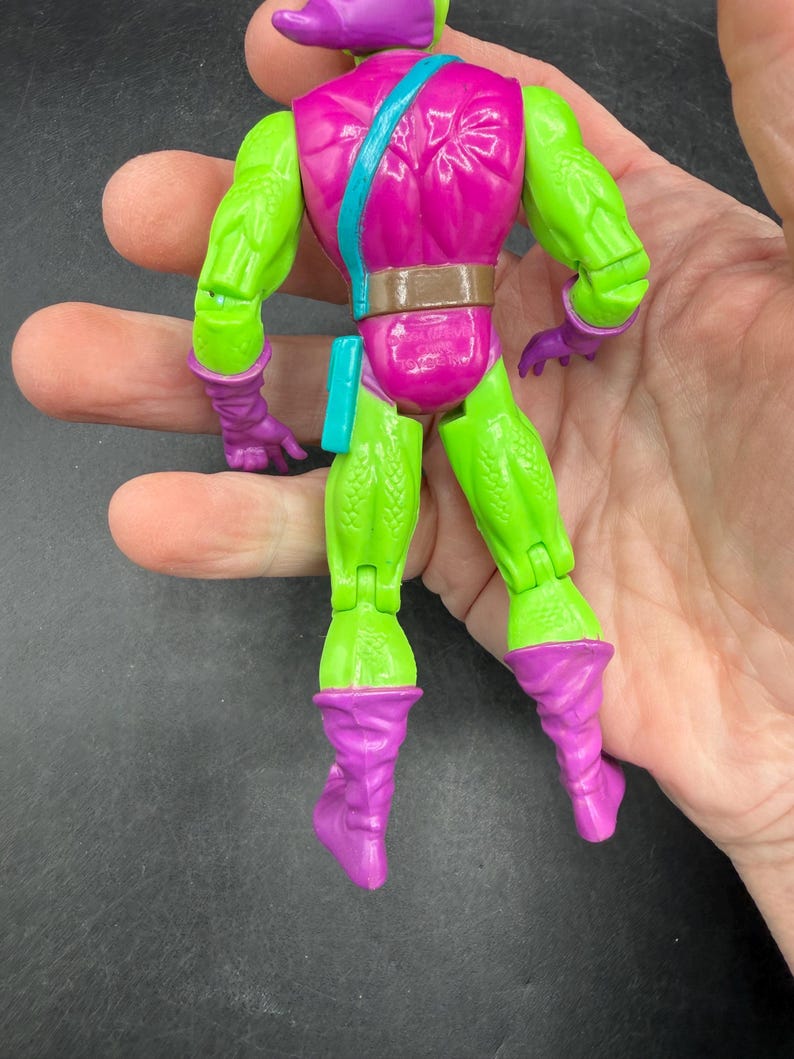 May include: A Green Goblin action figure with a purple head, gloves, and boots. The figure has a pink torso with a brown belt and a blue strap. The figure is held in a hand.