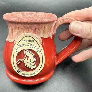 May include: A red ceramic mug with a glossy finish and a cream-colored drip glaze at the rim. A beige oval emblem on the front features the text Another Broken Egg Cafe, Hilton Head, SC, and a graphic of a cracked egg with a small turtle icon.