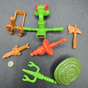 Puede incluir: Un conjunto de siete figuras de acción de las Tortugas Ninja Mutantes Adolescentes de plástico. Las figuras son verdes, naranjas, rojas, marrones y negras. Las figuras sostienen varias armas, incluyendo una espada, una lanza, un nunchaku, un sai y una katana.