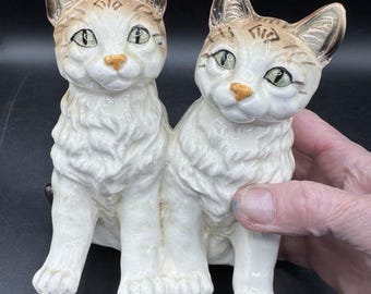 May include: A pair of ceramic cat figurines, sitting side-by-side. The cats are white with brown markings and have green eyes.