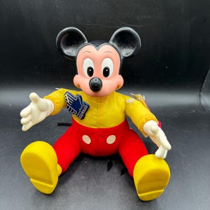 May include: A vintage Mickey Mouse plush doll. The doll features a yellow shirt, red pants, and large yellow shoes. It has black ears, a pink face, and white gloves. A blue tag with white text is attached to the doll.
