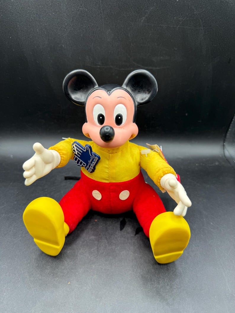 May include: A vintage Mickey Mouse plush doll. The doll features a yellow shirt, red pants, and large yellow shoes. It has black ears, a pink face, and white gloves. A blue tag with white text is attached to the doll.