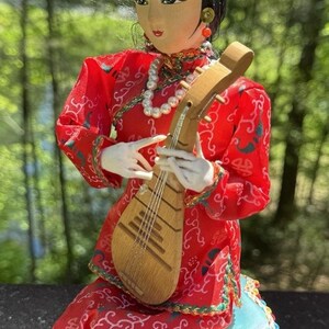 May include: A decorative doll in traditional Chinese attire, holding a wooden pipa. The doll wears a red dress with ornate patterns, a pearl necklace, and a gold headdress. It sits on a blue cushion with a decorative trim.