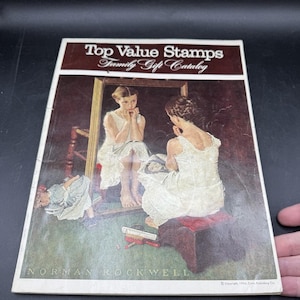 Puede incluir: Una portada vintage del catálogo de regalos para la familia Top Value Stamps con una ilustración de Norman Rockwell de dos chicas con vestidos blancos, una mirando en un espejo, con una muñeca en el suelo. El texto de la portada dice "Top Value Stamps Family Gift Catalog" y "Norman Rockwell".