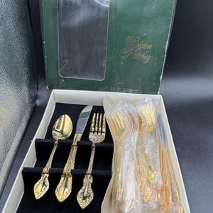 May include: A set of gold-toned cutlery in a green box labelled Golden Matchez. The set includes a spoon, a knife, and a fork with ornate, embossed handles. Some pieces are displayed in a black velvet tray, while others remain wrapped in plastic.