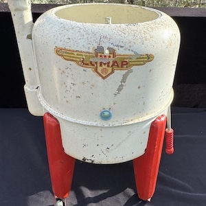 May include: Vintage white metal Lumar washing machine with red legs and wheels. The machine has a round top with a white and gold logo that says "Lumar" with three stars above the name.
