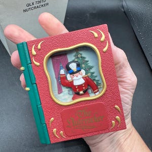 May include: A red and green decorative book-shaped ornament featuring a Nutcracker soldier in a framed scene. The ornament has gold accents and the text "The Nutcracker" on the front. The box reads "QLX 7261D NUTCRACKER".