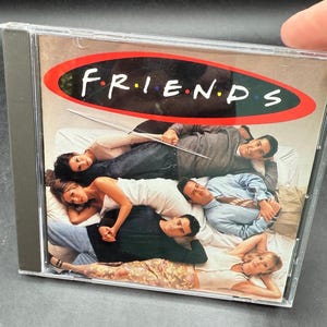 May include: A Friends CD case featuring the show's logo in white letters on a red oval. The cover image shows the six main characters lying on a bed. The CD case is in a clear plastic jewel case.