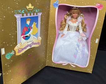 May include: A Disney Sleeping Beauty doll in a gold display box. The doll has long blonde hair and wears a white wedding gown with iridescent accents. The box features an illustration of the characters and the text Walt Disney's Sleeping Beauty.