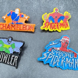 Może przedstawiać: Cztery kolorowe przypinki z postaciami z komiks&oacute;w. Przypinki zawierają Chameleona, Spider-Mana, The Prowler i Cyborg Spider-Man. Każda przypinka ma unikalny design i schemat kolor&oacute;w, z tekstem identyfikującym każdą postać.