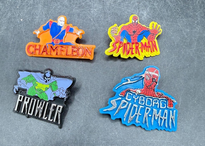Może przedstawiać: Cztery kolorowe przypinki z postaciami z komiks&oacute;w. Przypinki zawierają Chameleona, Spider-Mana, The Prowler i Cyborg Spider-Man. Każda przypinka ma unikalny design i schemat kolor&oacute;w, z tekstem identyfikującym każdą postać.
