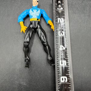 May include: A plastic action figure of a superhero, featuring a blue top with a white emblem, black pants, and yellow gloves. The figure is approximately 6 inches tall, measured with a black and white measuring tape.