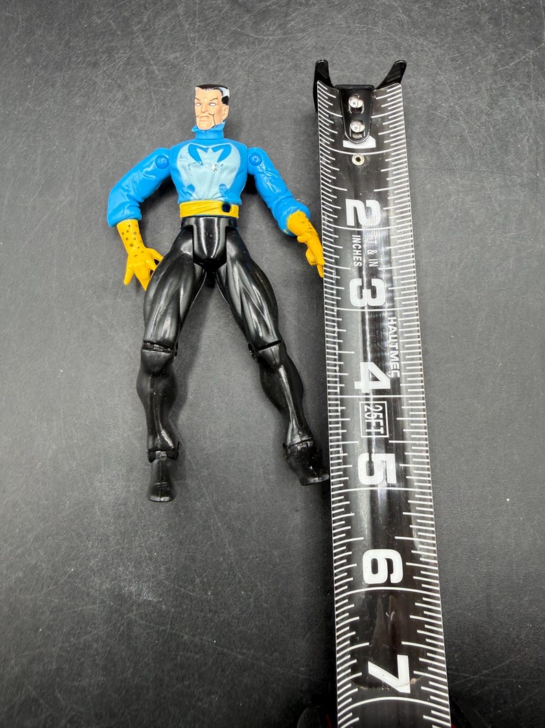 May include: A plastic action figure of a superhero, featuring a blue top with a white emblem, black pants, and yellow gloves. The figure is approximately 6 inches tall, measured with a black and white measuring tape.