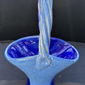 May include: A ceramic basket in shades of blue, featuring a twisted handle. The basket has a flared design, a rounded base, and a glossy finish. The handle has gold accents and is a lighter blue.