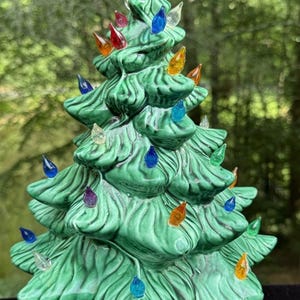 Peut inclure: Un sapin de Noël en céramique verte avec une finition brillante et des branches texturées. L'arbre est décoré de nombreuses petites ampoules en plastique translucide et multicolore, dans des tons bleu, rouge, orange, jaune, violet et transparent.