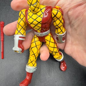 以下が含まれることがあります： 黄色と赤のアクションフィギュア。ダイヤモンド模様のデザイン。赤いブーツ、手袋、ヘッドカバーが付いています。手首、足首、ウエストにはシルバーのアクセント。左側には赤いアクセサリーがあります。