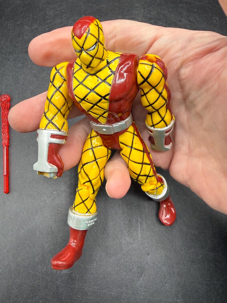 以下が含まれることがあります： 黄色と赤のアクションフィギュア。ダイヤモンド模様のデザイン。赤いブーツ、手袋、ヘッドカバーが付いています。手首、足首、ウエストにはシルバーのアクセント。左側には赤いアクセサリーがあります。