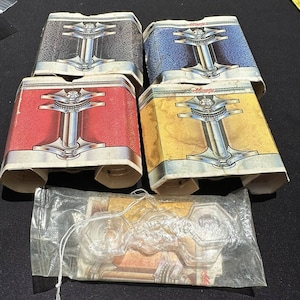 May include: Four vintage cardboard boxes with a retro design, each featuring a different color: black, blue, red, and yellow. Each box has a silver metal design in the center. The boxes are stacked on top of each other.