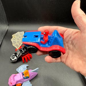 May include: A selection of colourful toy vehicles featuring Spider-Man and other characters. The Spider-Man car is red and blue, with a Spider-Man figure in the driver's seat. Other vehicles include a black and red spider-themed vehicle and a purple and blue airplane.