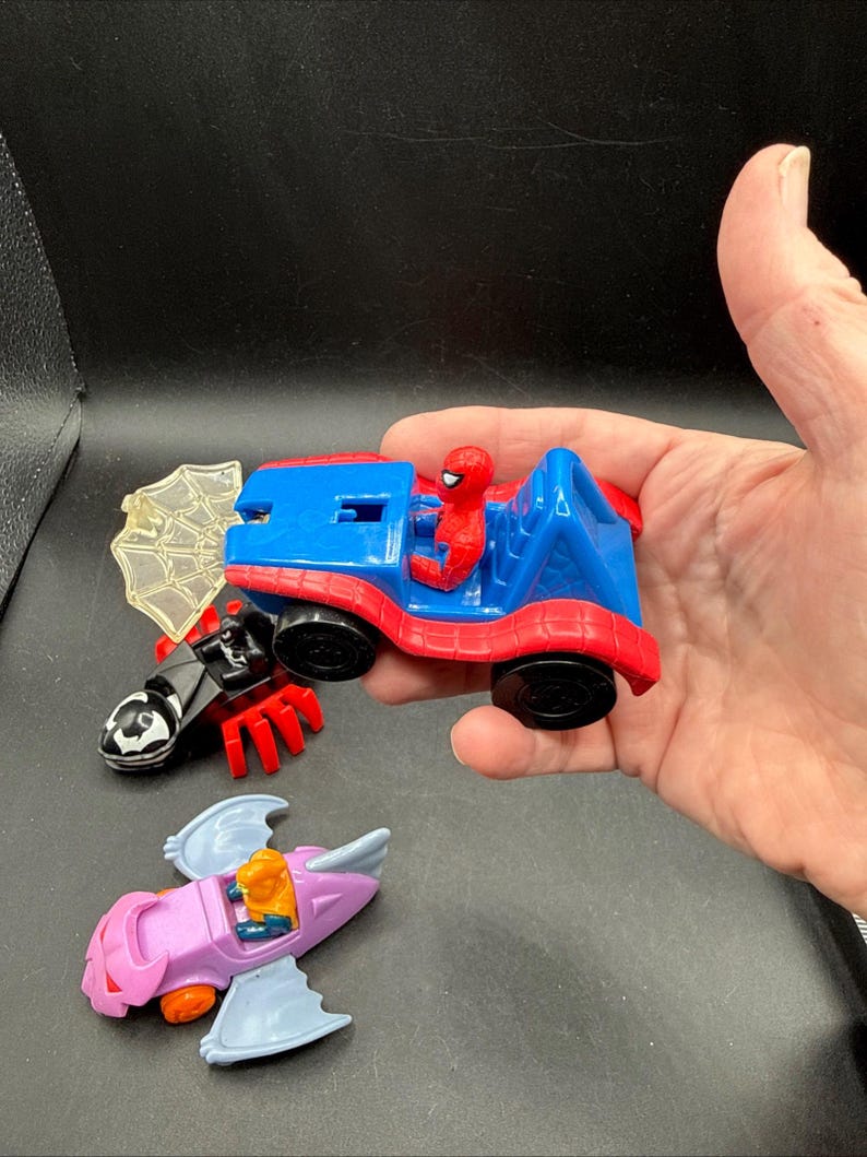 May include: A selection of colourful toy vehicles featuring Spider-Man and other characters. The Spider-Man car is red and blue, with a Spider-Man figure in the driver's seat. Other vehicles include a black and red spider-themed vehicle and a purple and blue airplane.