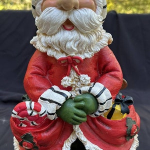 Puede incluir: Una figura de Papá Noel sonriente con barba blanca y mangas a rayas rojas y blancas. Lleva un bastón de caramelo y una bolsa de regalo en sus manos. Lleva un traje rojo con ribete blanco y un gorro a rayas blancas y negras.