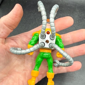 Puede incluir: Figura de acci&oacute;n del Dr. Octopus, vista desde atr&aacute;s, con un traje verde y amarillo y brazos mec&aacute;nicos plateados. La figura tiene botas naranjas y una pieza trasera de metal plateado.