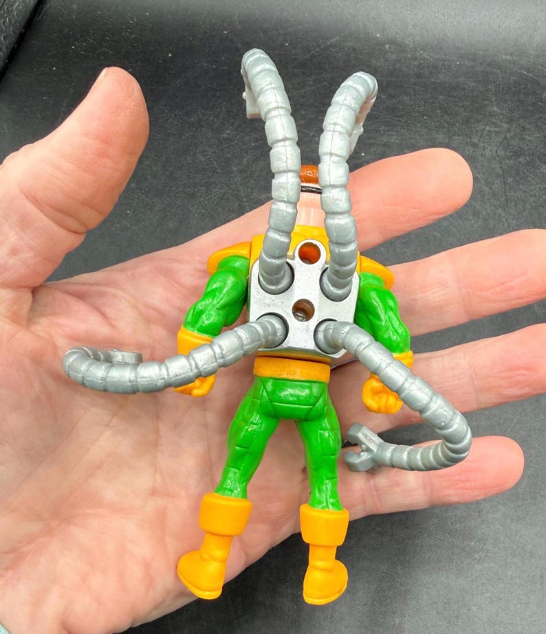 Puede incluir: Figura de acci&oacute;n del Dr. Octopus, vista desde atr&aacute;s, con un traje verde y amarillo y brazos mec&aacute;nicos plateados. La figura tiene botas naranjas y una pieza trasera de metal plateado.