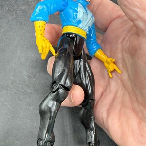 May include: A Dr. Strange action figure. The figure is wearing a blue jacket, yellow gloves, black pants, and a yellow belt. The figure has white hair and a stern expression. The figure is approximately 6 inches tall.