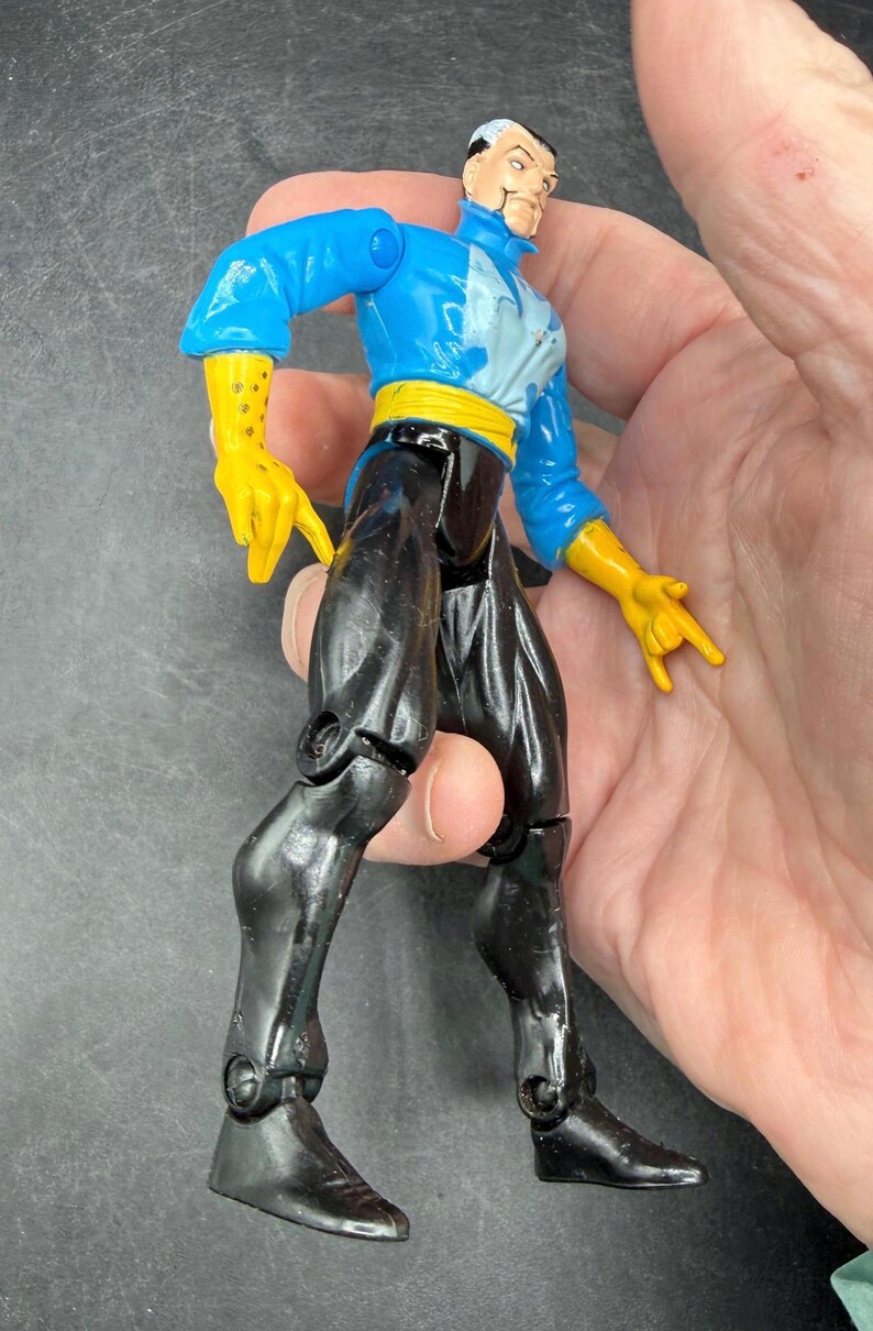 May include: A Dr. Strange action figure. The figure is wearing a blue jacket, yellow gloves, black pants, and a yellow belt. The figure has white hair and a stern expression. The figure is approximately 6 inches tall.