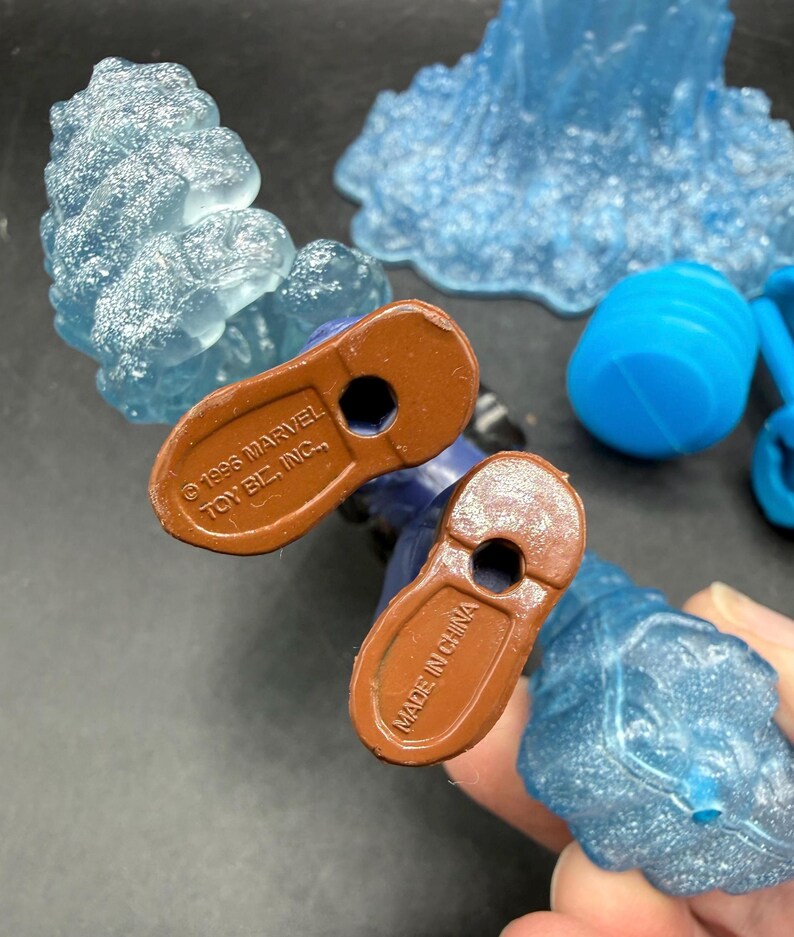 May include: A close-up shot of a blue action figure's feet, with brown soles. The soles have the text "&copy; 1995 MARVEL TOY B.L., INC." and "MADE IN CHINA". Several translucent blue ice-like accessories are scattered around.