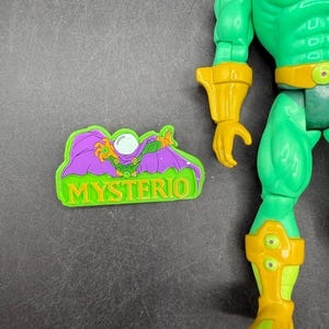 Op de afbeelding: Een groen en geel actiefiguur van Mysterio, een stripboekpersonage, met een bijpassende magneet. Het figuur heeft een groene body, gele accenten en een paarse helm. De magneet bevat het woord "MYSTERIO".