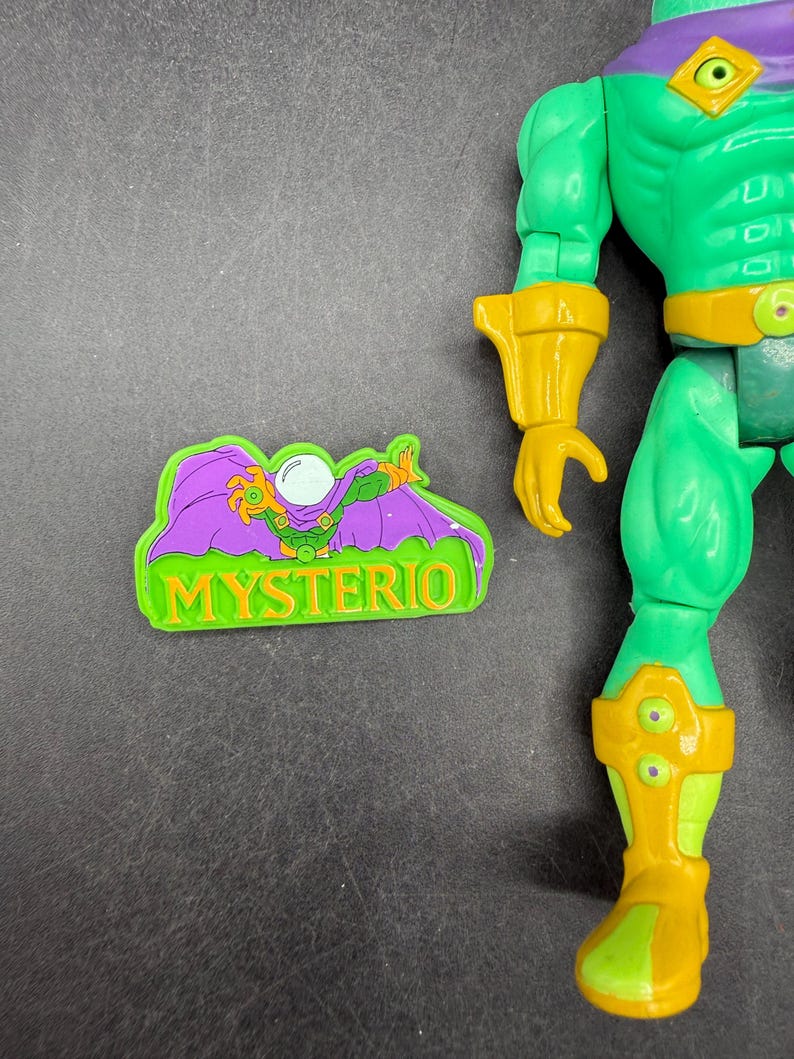 Op de afbeelding: Een groen en geel actiefiguur van Mysterio, een stripboekpersonage, met een bijpassende magneet. Het figuur heeft een groene body, gele accenten en een paarse helm. De magneet bevat het woord "MYSTERIO".