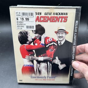 May include: A DVD case for the film The Replacements featuring Keanu Reeves and Gene Hackman. The cover art displays a football player in a red jersey, a cheerleader in a red outfit, and a man in a suit and straw hat. Text includes the title, actor names, and the quote Enormously Funny.