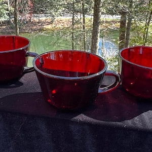 May include: Three ruby red glass mugs with black handles. The mugs are angled and have a glossy finish. The mugs are sitting on a black surface with a natural background.