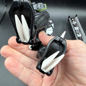 May include: A black and white action figure with large white claws. The figure has a detailed design with a green accent on its back. A ruler is in the background.