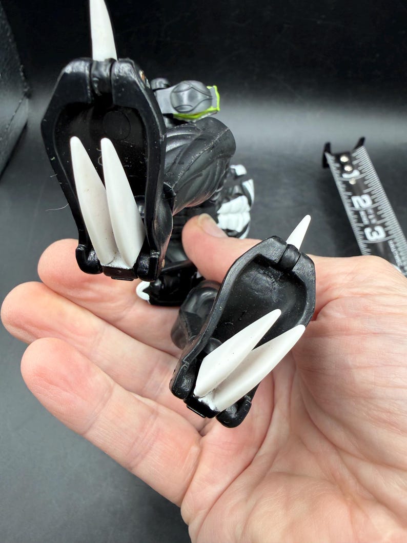 May include: A black and white action figure with large white claws. The figure has a detailed design with a green accent on its back. A ruler is in the background.