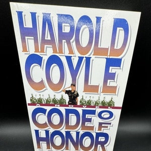 Op de afbeelding: Een witte boekomslag met de titel "Code of Honor" in blauwe en oranje letters. De naam van de auteur, "Harold Coyle", is in blauwe en oranje letters boven de titel gedrukt. Een vrouw in militair uniform staat voor een rij soldaten en salueert.