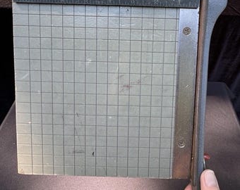 May include: Vintage paper cutter by Milton Bradley Company. The gray cutting surface features a grid pattern and a metal ruler along the top edge. The cutter has a gray handle and blade, with the brand name and patent information visible.