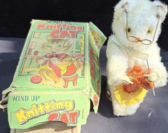May include: A vintage wind-up toy featuring a white plush cat wearing glasses and knitting with orange yarn. The toy is next to its original box, which has a green background and the words "Knitting Cat" in yellow and red.