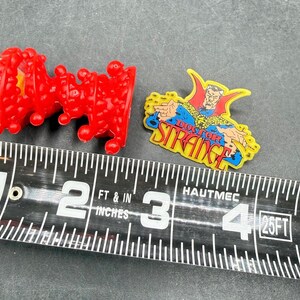 K&ouml;nnte beinhalten: Zwei Marvel-Pins und ein rotes Plastikspielzeug werden neben einem Ma&szlig;band angezeigt. Ein Pin zeigt Doctor Strange mit dem Text "Doctor Strange". Das andere ist ein rotes, strukturiertes Plastikspielzeug. Das Ma&szlig;band zeigt Zentimeter.