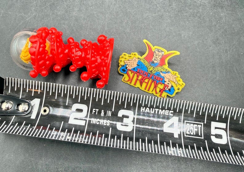 K&ouml;nnte beinhalten: Zwei Marvel-Pins und ein rotes Plastikspielzeug werden neben einem Ma&szlig;band angezeigt. Ein Pin zeigt Doctor Strange mit dem Text "Doctor Strange". Das andere ist ein rotes, strukturiertes Plastikspielzeug. Das Ma&szlig;band zeigt Zentimeter.