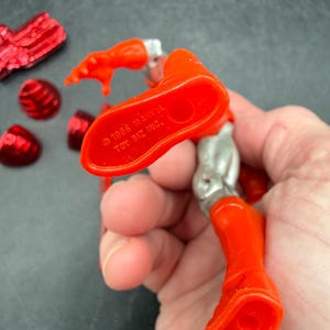 Puede incluir: Una figura de acci&oacute;n roja y plateada con el texto "&copy; 1995 MARVEL TOY BIZ INC." y "MADE IN CHINA" en el pie. Varios accesorios rojos est&aacute;n esparcidos alrededor de la figura.