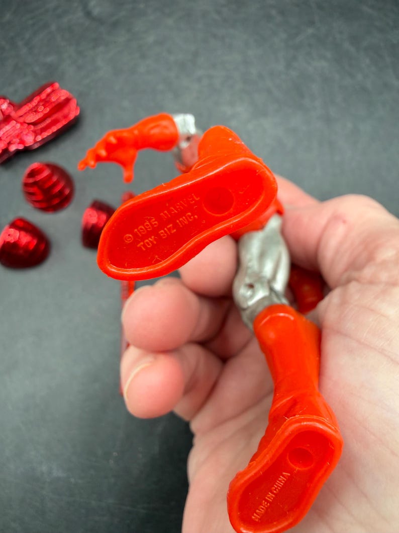 Puede incluir: Una figura de acci&oacute;n roja y plateada con el texto "&copy; 1995 MARVEL TOY BIZ INC." y "MADE IN CHINA" en el pie. Varios accesorios rojos est&aacute;n esparcidos alrededor de la figura.