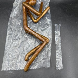 May include: A bronze-colored metal sculpture of a person in a seated position, packaged in a clear plastic bag. The sculpture has a minimalist design with elongated limbs and a rounded head. The figure is approximately 10 inches tall.