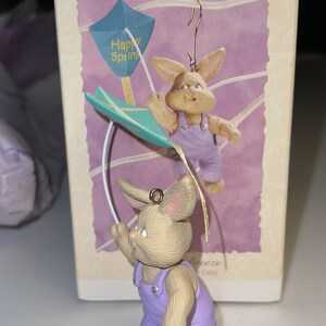 May include: A Hallmark Keepsake ornament featuring two bunny figurines. The bunnies wear purple dungarees and hold a teal kite with "Happy Spring" printed on it. The ornament is in its original packaging.