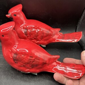 Könnte beinhalten: Zwei Keramik-Kardinalvogel-Figuren in leuchtendem Rot. Die Vögel haben detaillierte Flügel und Schwanzfedern mit schwarzen Akzenten an Schnäbeln und Augen. Die Figuren sind nebeneinander positioniert und zeigen ihre glänzende Oberfläche und dekorative Ausstrahlung.