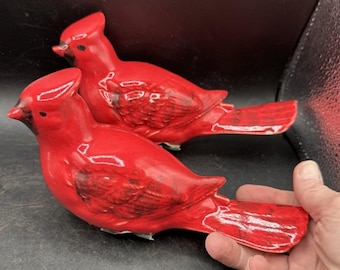 以下が含まれることがあります： 鮮やかな赤色のセラミックカーディナルバードの置物2つ。鳥は詳細な翼と尾羽を持ち、くちばしと目に黒いアクセントがあります。置物は並んで配置されており、光沢のある仕上がりと装飾的な魅力を示しています。