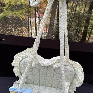 May include: A light green floral print baby carrier with blue bows and yellow wooden accents. The carrier has a ruffled edge and padded interior, with matching floral print straps.