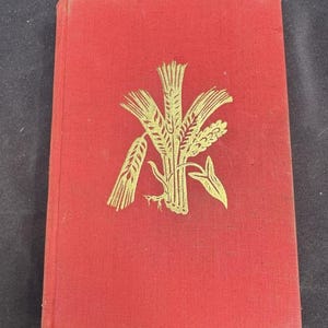 May include: A red hardcover book with a gold wheat sheaf design on the cover. The book has a textured surface and appears to be vintage. The cover design features stylized wheat stalks and leaves.